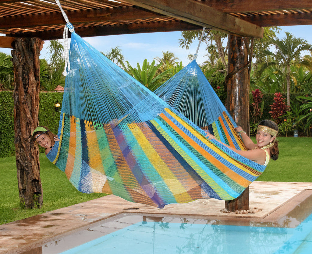 Mexi 4 Maya-Hängematte in bunten Farben über einem Pool, darin liegen zwei Mädchen leicht aufrecht quer, die Füße einander zugewandt und halten die Hängematte dadurch offen.