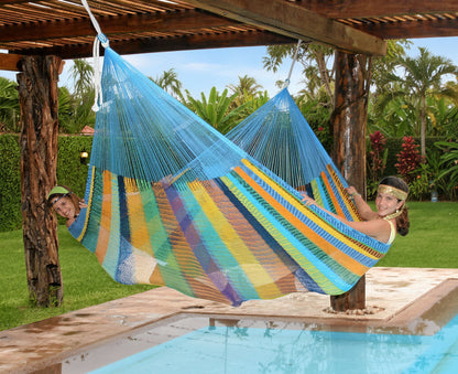 Mexi 4 Maya-Hängematte in bunten Farben über einem Pool, darin liegen zwei Mädchen leicht aufrecht quer, die Füße einander zugewandt und halten die Hängematte dadurch offen.